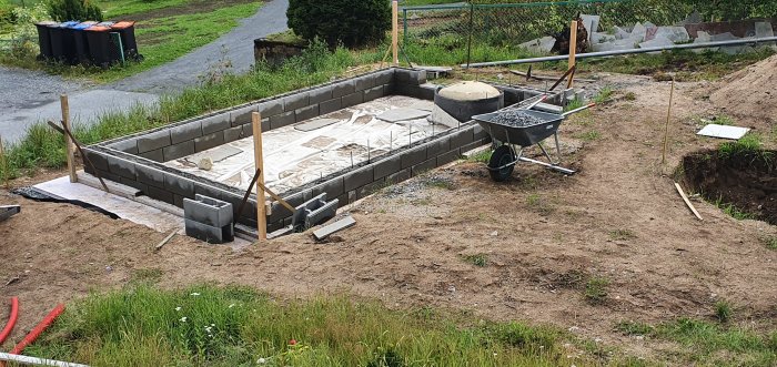 Pågående konstruktion av ett växthus med en ringmur av cementblock och isolerplattor på markduken, grävt område syns.