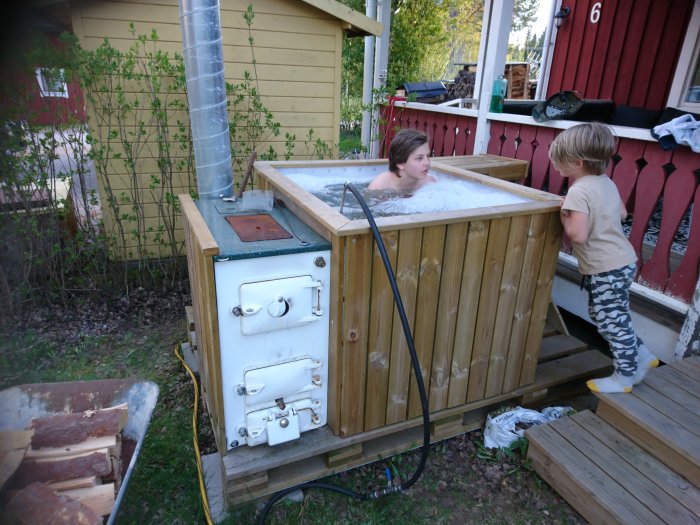 Två barn vid en hemmagjord badtunna med vedeldad panna utomhus nära en röd stuga.