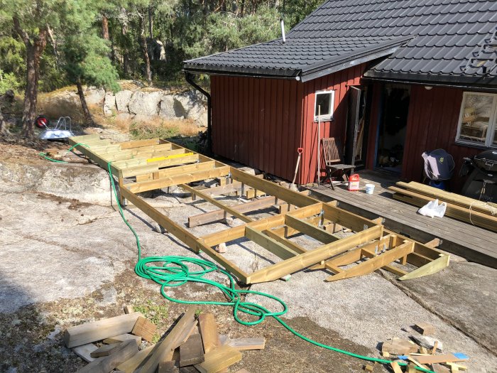 Träreglar som utgör grunden för en konstruktion vid en röd stuga, med betonggjutning runt stolpskor.