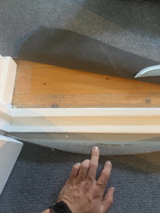 En hand som lyfter en mörkgrå matta för att blotta ett underliggande trägolv vid en dörröppning.