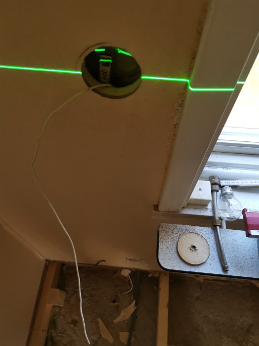 Laserlinje markerar installationshöjd för eluttag vid renoveringsprojekt, uttagshål i vägg med dragtråd och verktyg synliga.