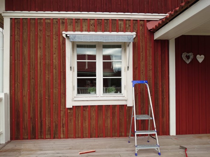 Rödmålad träfasad med vita fönsterkarmar och nylagt lockläkt, och en blå stege framför på en trädäck.