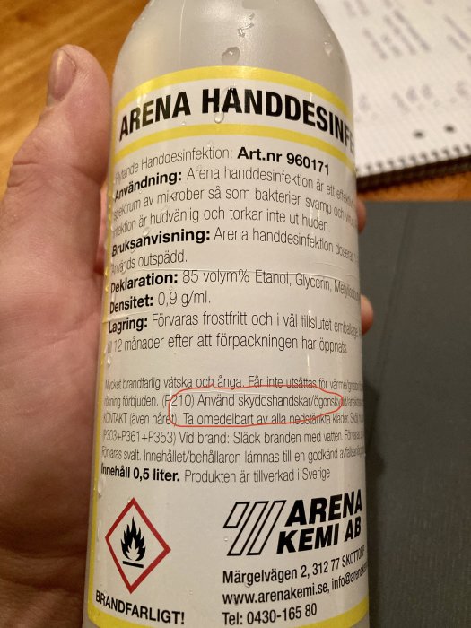 Hand håller en flaska med texten "ARENA HÄNDDESINFEKTION", säkerhetsvarningar och bruksanvisning synlig.