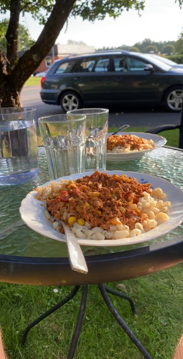 Måltid med pasta och köttfärssås serverad på ett utomhusbord med glas vatten, med suddig bakgrund av bil och träd.