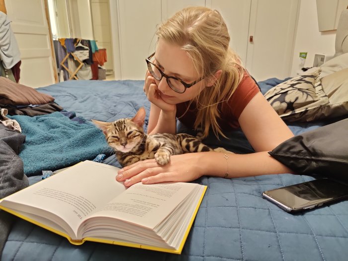 Kvinna som försöker läsa en bok med en sovande katt på armen, ligger på en blå täcke.