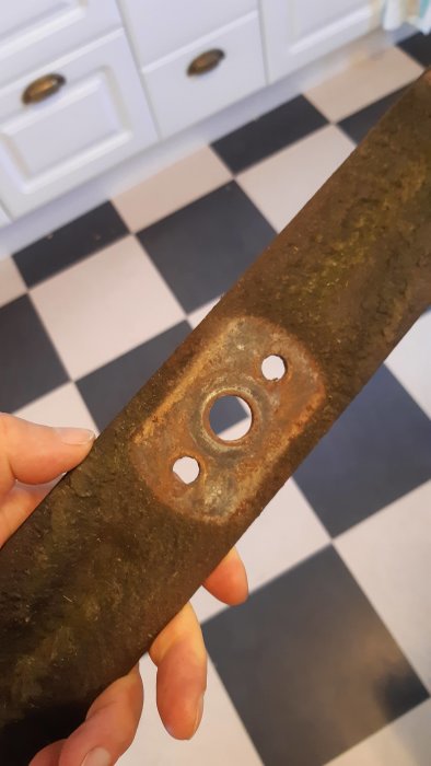 Hand håller en rostig metallinfästning med ett 16 mm centralt hål och två 9,5 mm hål, framför svartvit rutigt golv.