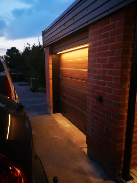 Varmgul LEDstrip ovanför en stängd garageport vid skymning, ger ett mjukt ljus mot garagets tegelvägg och port.