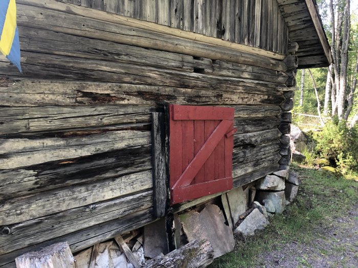 Äldre timmrad lada med skadade trästockar och en röd dörr, behöver renoveras.