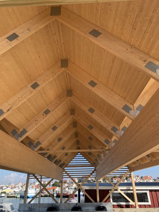 Träkonstruktion av en carport med öppet råsponttak och synliga bjälkar mot blå himmel och kuststad i bakgrunden.