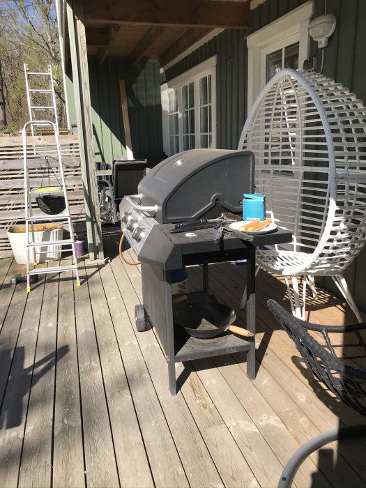Trädäck under renovering med grill, trädgårdsmöbler och en stege i bakgrunden.