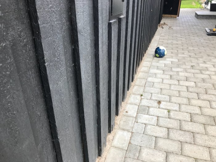 Svart träpanel möter grå markstenar på gården, skador vid kontaktområde och en fotboll syns i hörnet.