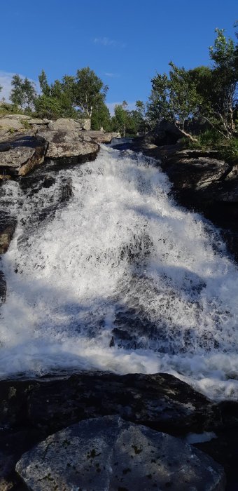 Vattenflödet i Römmesfossen med stenar och grönska runtomkring under klarblå himmel.