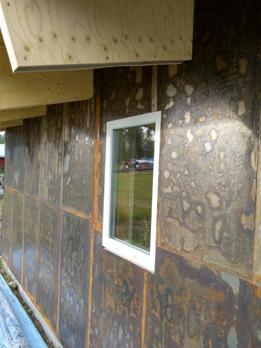 Rostiga cortenstålplåtar monterade runt fönster på hus, med synliga skruvar och överlappande skarvar.