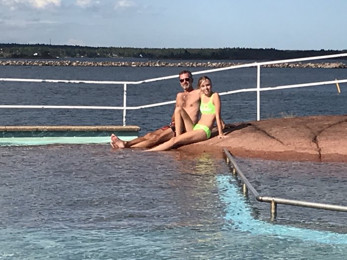 Två personer sitter avslappnat vid kanten av en pool som integrerats med naturliga berg, med havsutsikt i bakgrunden.