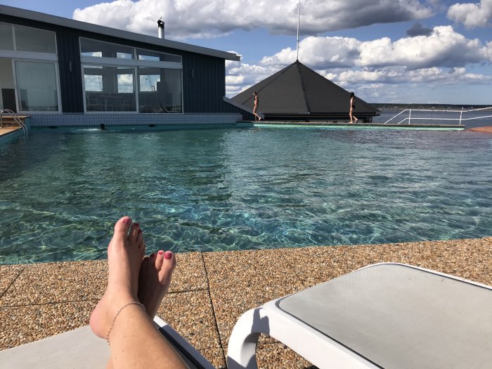 Vy över en klarblå pool från kanten med en persons fötter i förgrunden, och utsikt mot hav och horisont i bakgrunden.