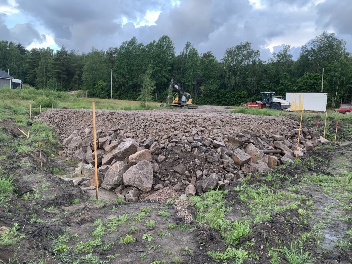 Byggplats med hög av grov grus och sten, angränsande skog, utsättningsstick och maskiner i bakgrunden.