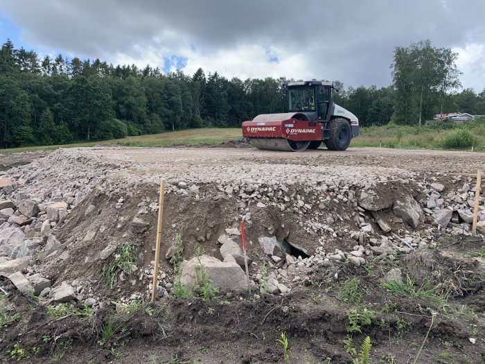 Vältmaskin jämnar ut ett lager av grus på en byggplats med skog i bakgrunden.