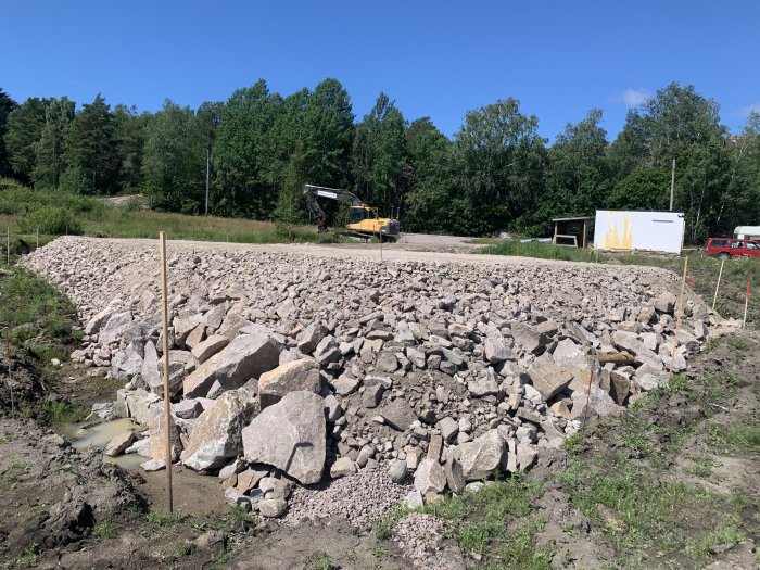 Byggplats med stora stenar, grus och markeringsspikar efter utsättning, grävmaskin i bakgrunden.