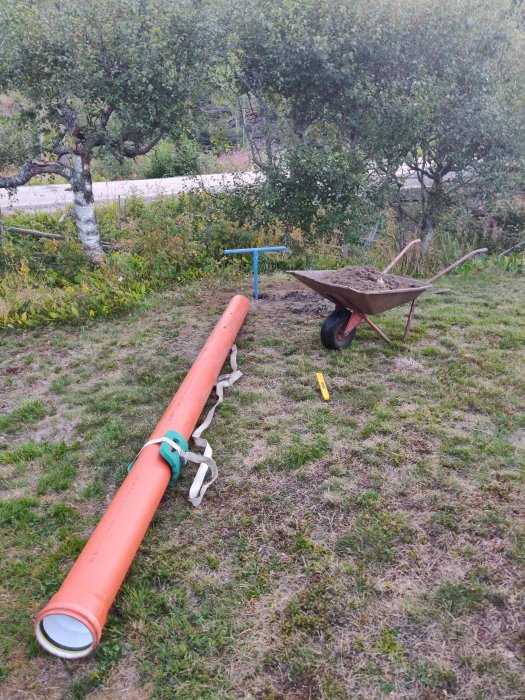 Långt orange foderrör på marken med hjulburen skottkärra full av sand i bakgrunden.