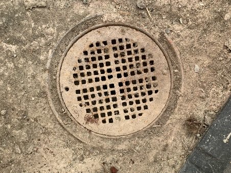 Gammal regnvattenbrunn med rutigt galler på marken som kan behöva tillfällig pluggning.