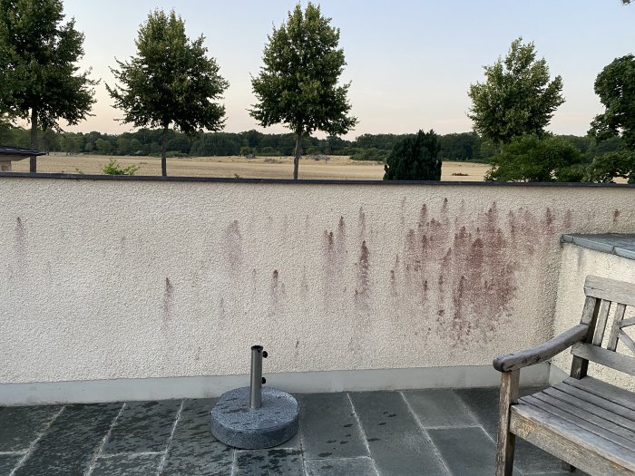 Vit sprutputsad vägg med röda utfällningar, terrassutsikt med träd i bakgrunden.