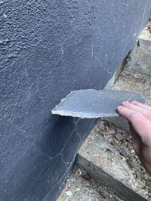 Hand håller en bit av lös puts för att visa dess tjocklek mot en väggyta.