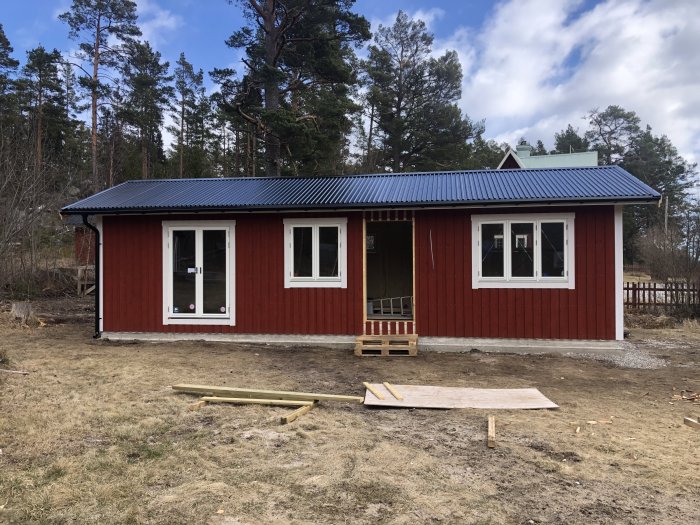 Röd trästuga med svart plåttak och vita fönsterkarmar, omgiven av skog och byggmaterial på marken.