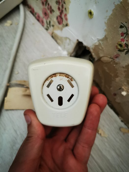 Hand håller ett gammalt telefonuttag märkt med "TELE", vid en öppning i väggen med synliga kablar.