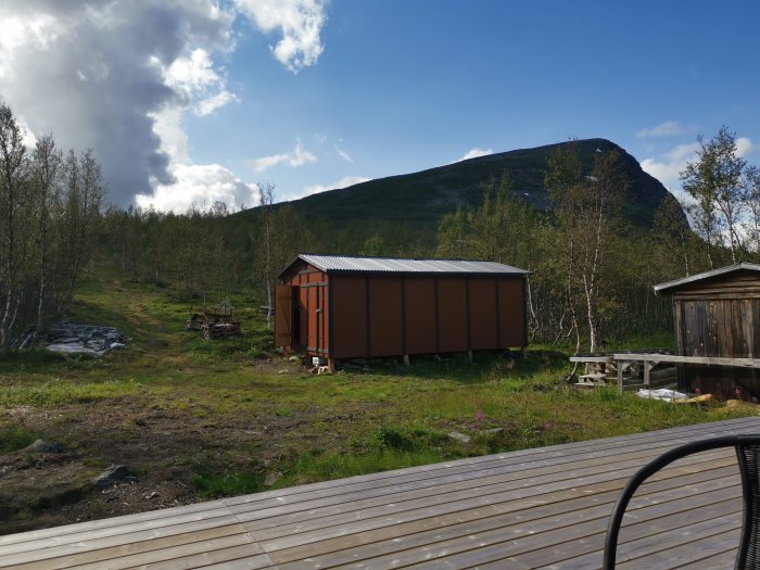 Stuga med lutande ben omgiven av natur med ett berg i bakgrunden.