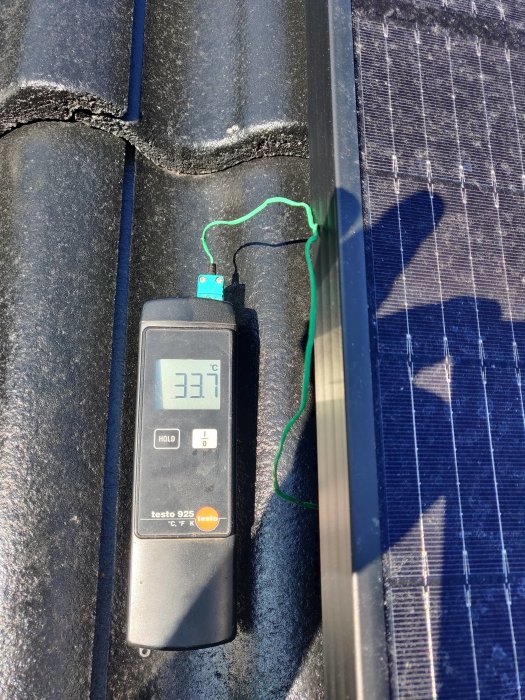 Termometer visar 33,7 °C på en svart takpanna vid sidan av ett solpanel.