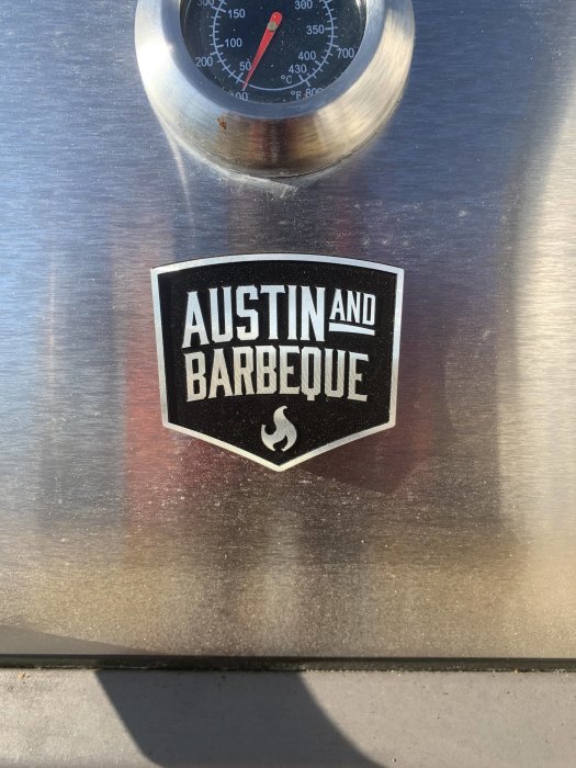 Termometer på grill med logotyp och text 'AUSTIN AND BARBEQUE' samt en låga-symbol.