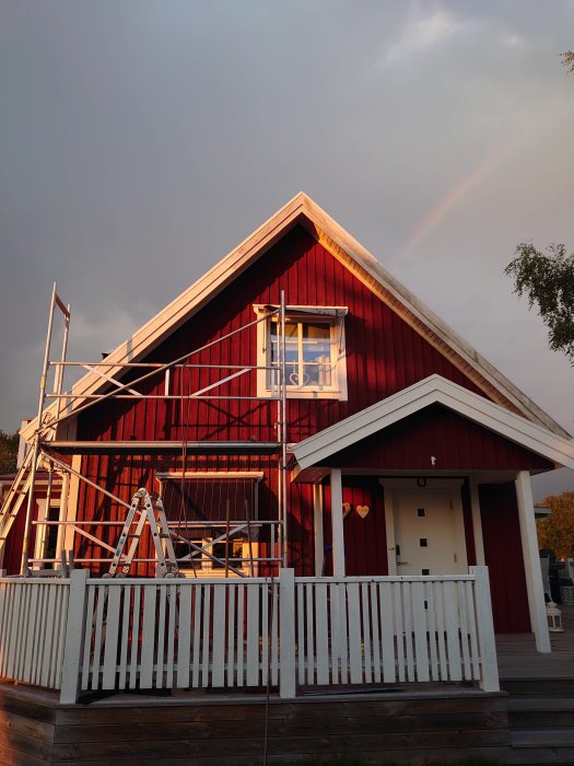 Rödmålat hus med delvis tvättad fasad och stege, regnbåge i bakgrunden.