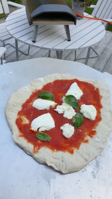 Ojämn hemlagad pizza före gräddning med tomatsås, mozzarella och basilika på utebord.