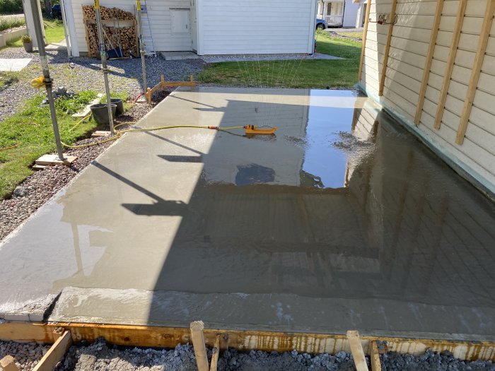 Nyutgjuten och vattenmättad betongplatta bredvid hus med reflekterande vattenpölar och skuggor.