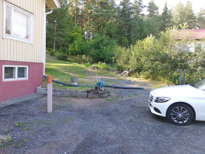 Hörn av ett hus med störtflod som leder regnvatten mot en ojämn gård och en parkerad bil.