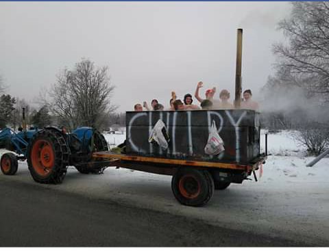 Människor badar i en mobil badtunna dragen av en traktor på vintern med snö i bakgrunden.