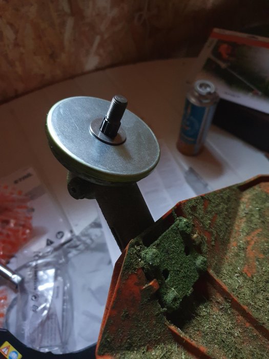 En roterande trimmerhuvud med en skruv i förgrunden och en delvis synlig trimmer med gräsrester i bakgrunden, på ett arbetsbord.