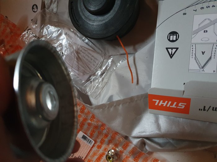 Hand håller en del av maskin nära kamera, med oöppnad förpackning och manual i bakgrunden.
