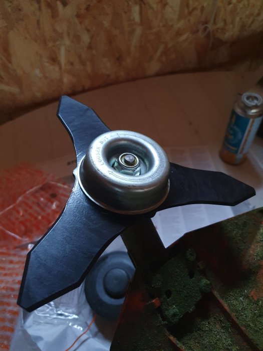 Närbild på en nyligen monterad grästrimmerkniv med fyra blad inne i ett verkstadsutrymme, med en tom läskburk och verktyg i bakgrunden.