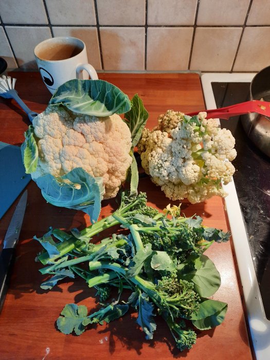Blomkålshuvuden och broccoligrenar på ett träköksbord bredvid en kniv och en kaffekopp.
