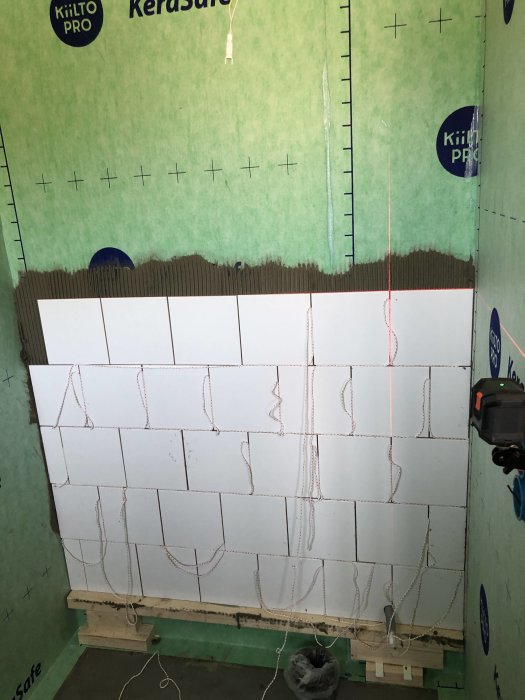 Halvpåbörjad kakling med vita plattor i halvförband mot grön tätskiktsvägg, överskott av fix och synligt rött snöre.