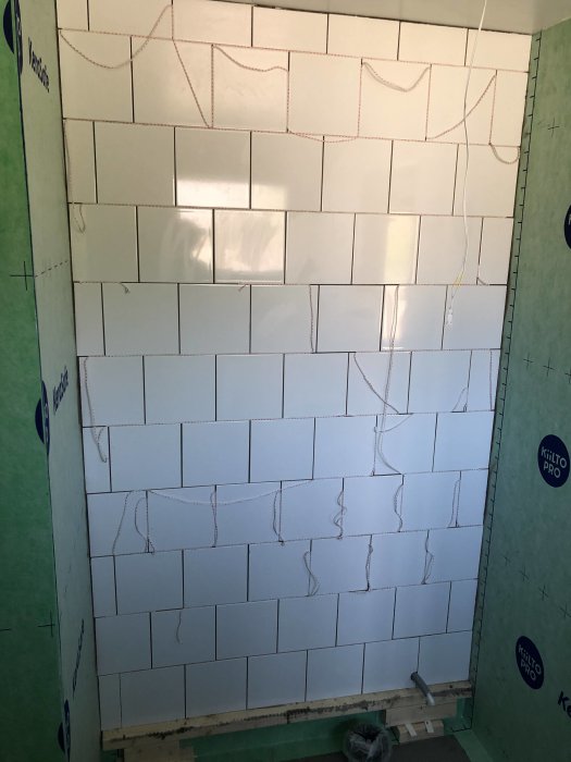 Pågående kakelarbete på en vägg med halvfärdigt vitt kakel i halvförband och synliga kakelsnören.
