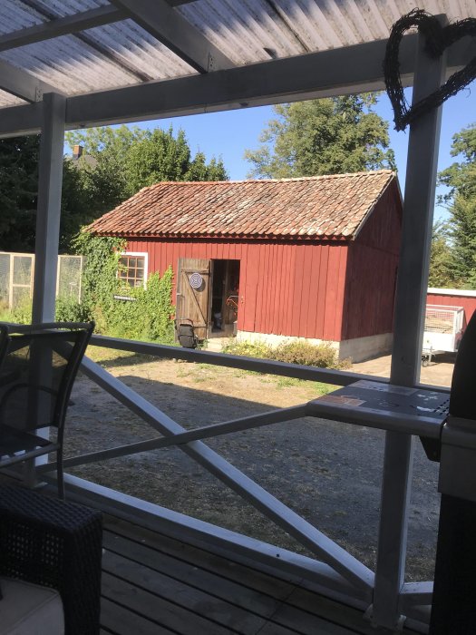 Röd träbod med tegeltak sedd från en veranda, omgiven av grönska och klarblå himmel.