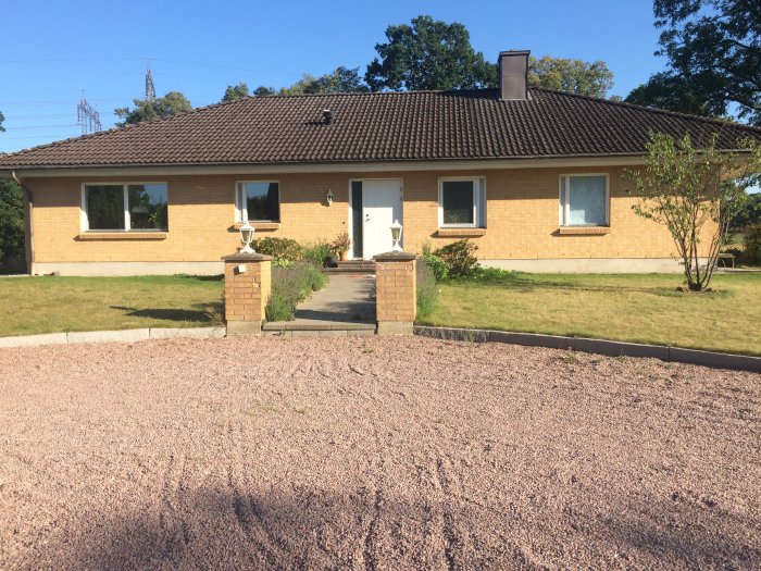 Ett gult tegelhus med brunt tak, vit entrédörr och vita fönster, grönt gräs och grusgång.