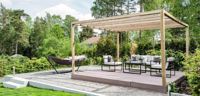 Modern pergola med trästolpar och balkar över en trallterrass med utemöbler och hängmatta, placerad i en trädgård.