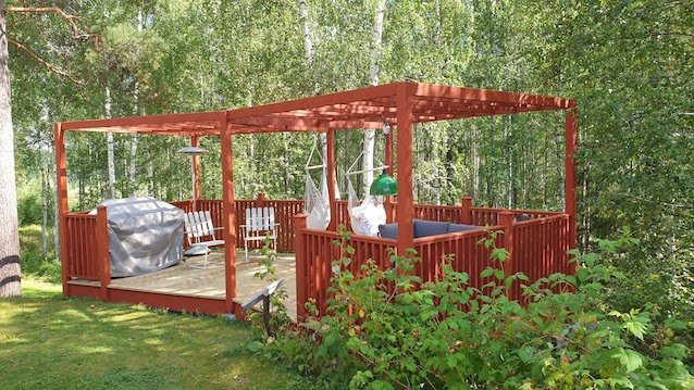 Nybyggd pergola med röda stolpar och staket, avfasad ram samt centraltakregel, omgiven av grönska.