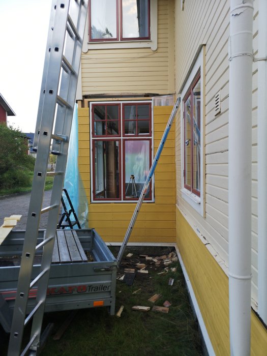 En pågående byggnadsrenovering med en stege mot en gul trävilla och en släpvagn fylld med träplankor.