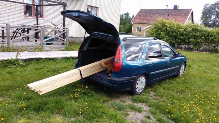 En personbil lastad med långa brädor som sticker ut från den öppna bakluckan, parkerad på en gräsmatta.