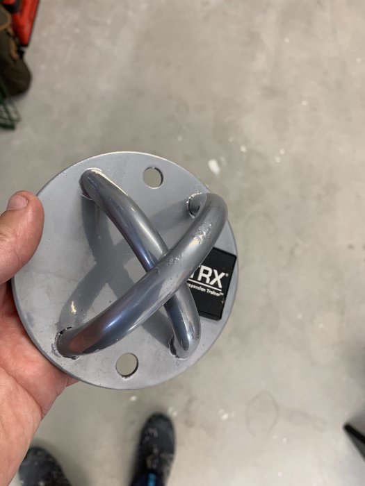 Metallkrok för träningsredskap med rund bas och fyra infästningshål, hållen nära golvet.