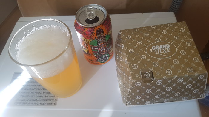 En öppnad ölburk och ett glas med öl bredvid en stängd take away låda med texten Grand de Luxe.